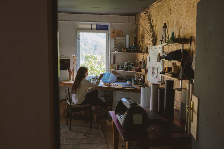 A woman artist painting at her work table in a cozy home studio with natural light.
