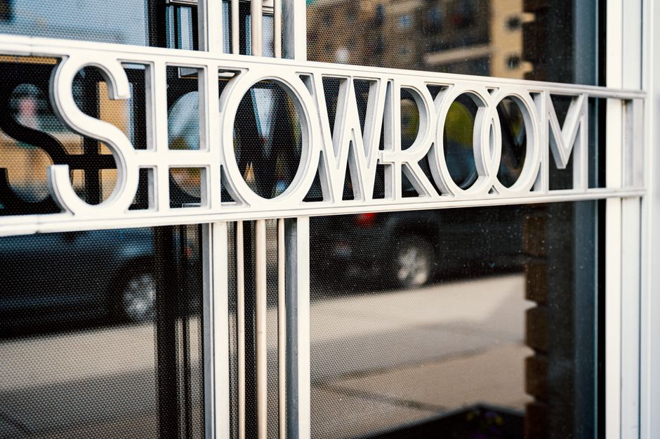 Store with Showroom title on glass window reflecting tiled pavement and road with cars behind multistage building in town in daylight