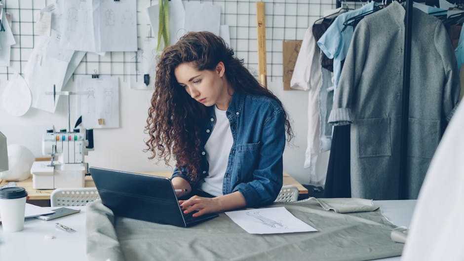 Young designer using laptop in modern studio surrounded by sketches and fabrics.